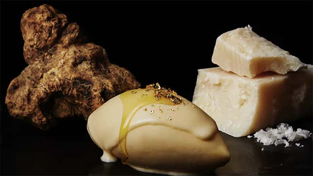 An almond-shaped, light-colored ice cream scoop with oil and gold flakes on top sits in front of a black background near a white truffle and Parmesan