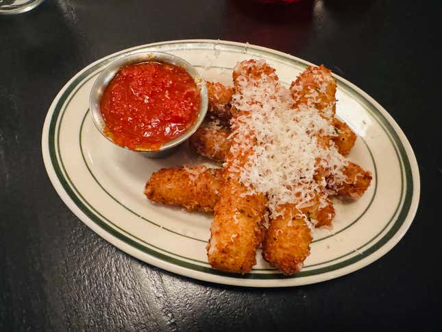 Mozzarella sticks dusted with cheese sit on a white plate on a black table with red dipping sauce