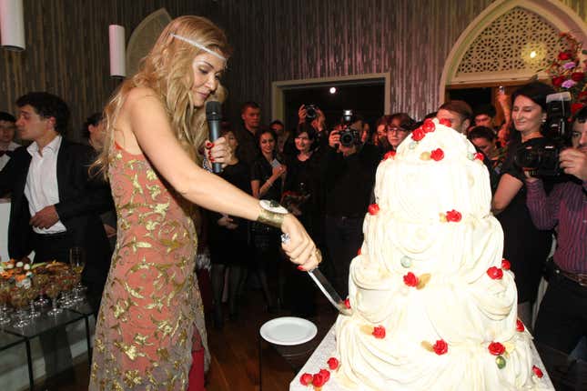 A blonde woman in a light pink and gold dress cuts a piece from an ornate white cake
