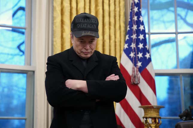 Elon Musk looks downcast while wearing all black in front of a U.S. flag in the Oval Office