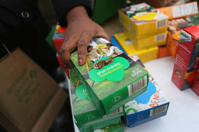 A hand holds three boxes of Thin Mint cookies on a table with boxes of lots of other types