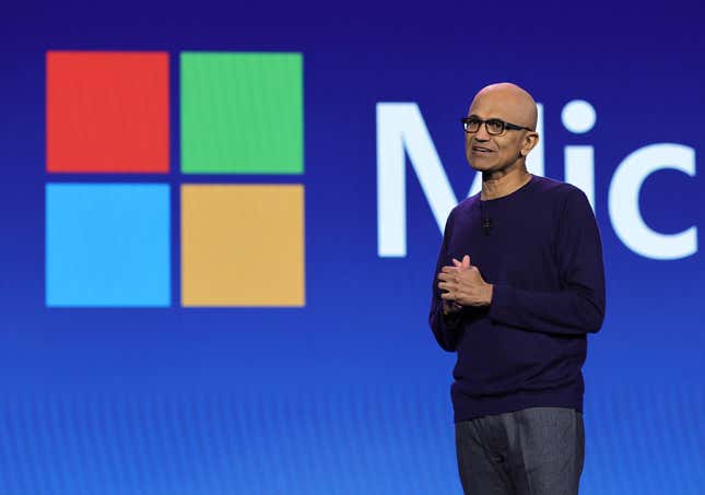 Satya Nadella speaking with his hands clasped together in front of a Microsoft logo on a blue backdrop, he's wearing a navy sweater and grey pants