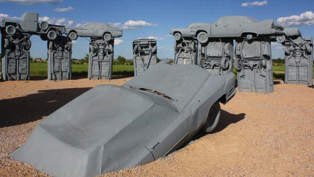 Gray cars staged to look like Stonehenge surround a car in the dirt; the sky behind is blue with a few clouds